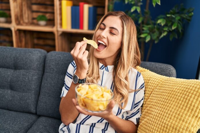 Young,Blonde,Woman,Eating,Chips,Potatoes,Sitting,On,Sofa,At